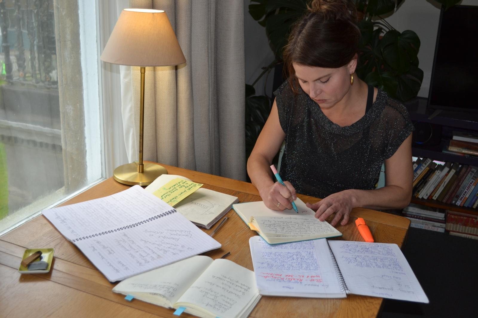 Elizabeth Welty at a wooden desk, writing in a notebook with several other notebooks open beside her, a brass lamp lit and a window to the left.
